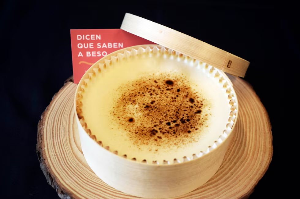 Una tarta de queso de La Lucastería en su molde de papel plisado sobre una tabla de madera, con una tarjeta roja apoyada detrás en la que se lee «Dicen que saben a beso».