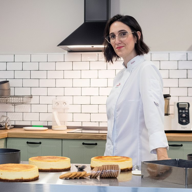 María Lucas, con chaquetilla blanca, de pie en el obrador de La Lucastería junto a la mesa de acero con las tartas de queso recién hechas.