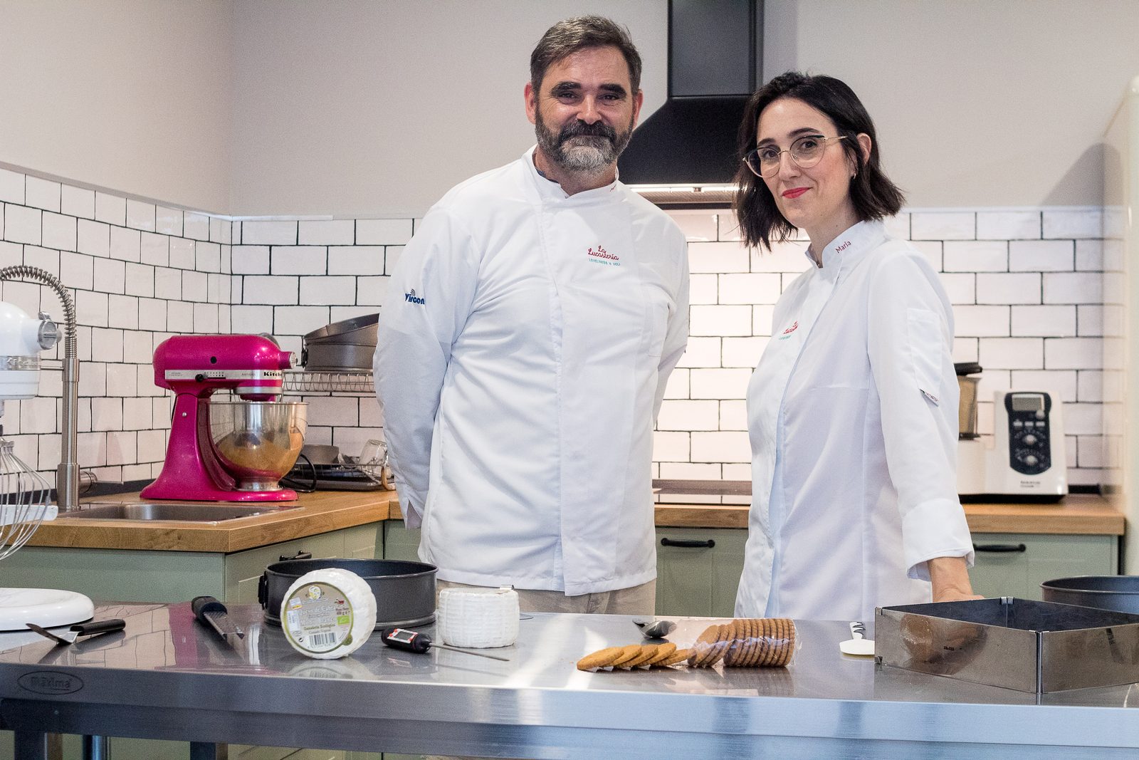 María Lucas y Miguel Ángel Mateos, con chaquetilla blanca, en el obrador de azulejo blanco de La Lucastería, junto a una mesa de acero con tartas de queso y una amasadora.