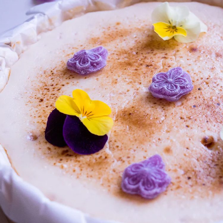 Tarta de queso de violetas de La Lucastería, decorada con rosetones de crema morada y flores comestibles amarillas y moradas.