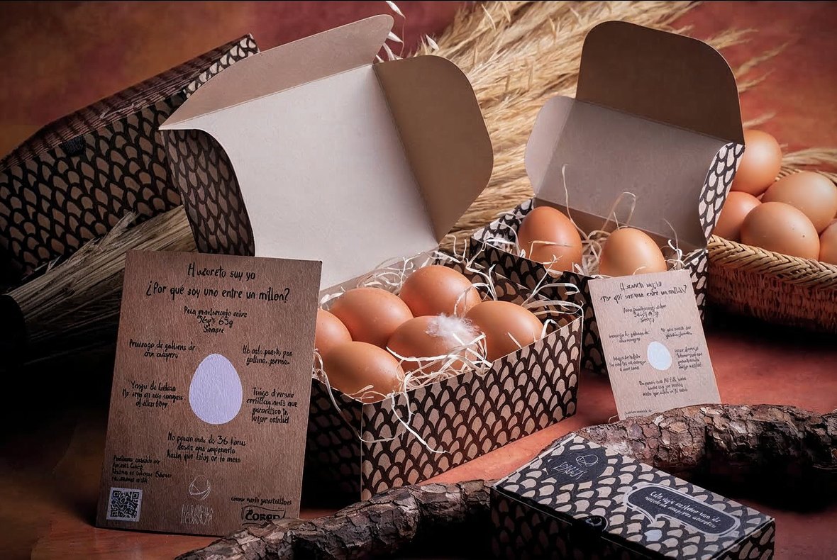 Las cajas de huevos de Taberna Pedraza, abiertas sobre paja y con el cartón estampado con un patrón de escamas. Al lado, la tarjeta escrita a rotulador que explica en primera persona por qué el huevo es «uno entre un millón», y una caja cerrada con el mensaje «Esta caja contiene uno de nuestros mayores secretos».