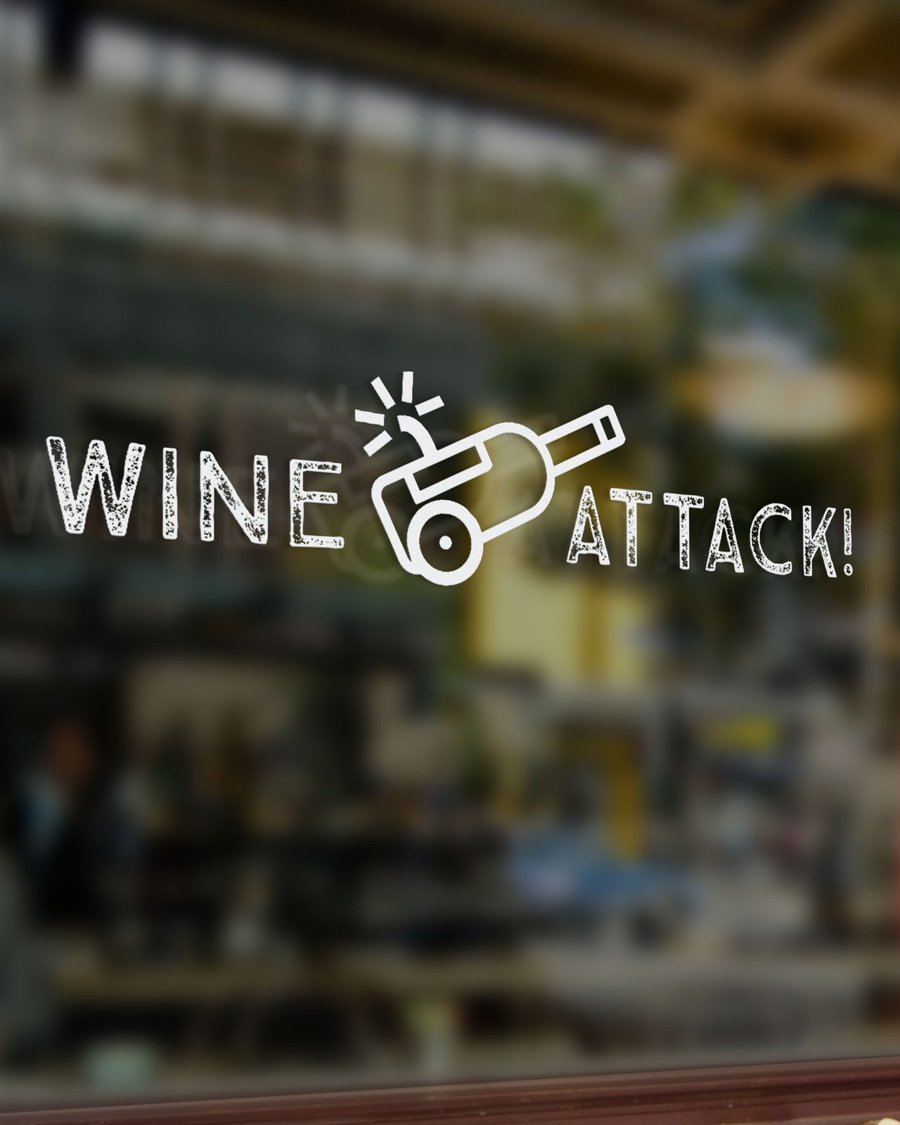 El logotipo de Wine Attack en vinilo blanco sobre el cristal del escaparate: las palabras WINE y ATTACK! separadas por una botella tumbada dibujada como un cañón, con la mecha encendida y el corcho saliendo disparado. Detrás se adivina el interior del local.